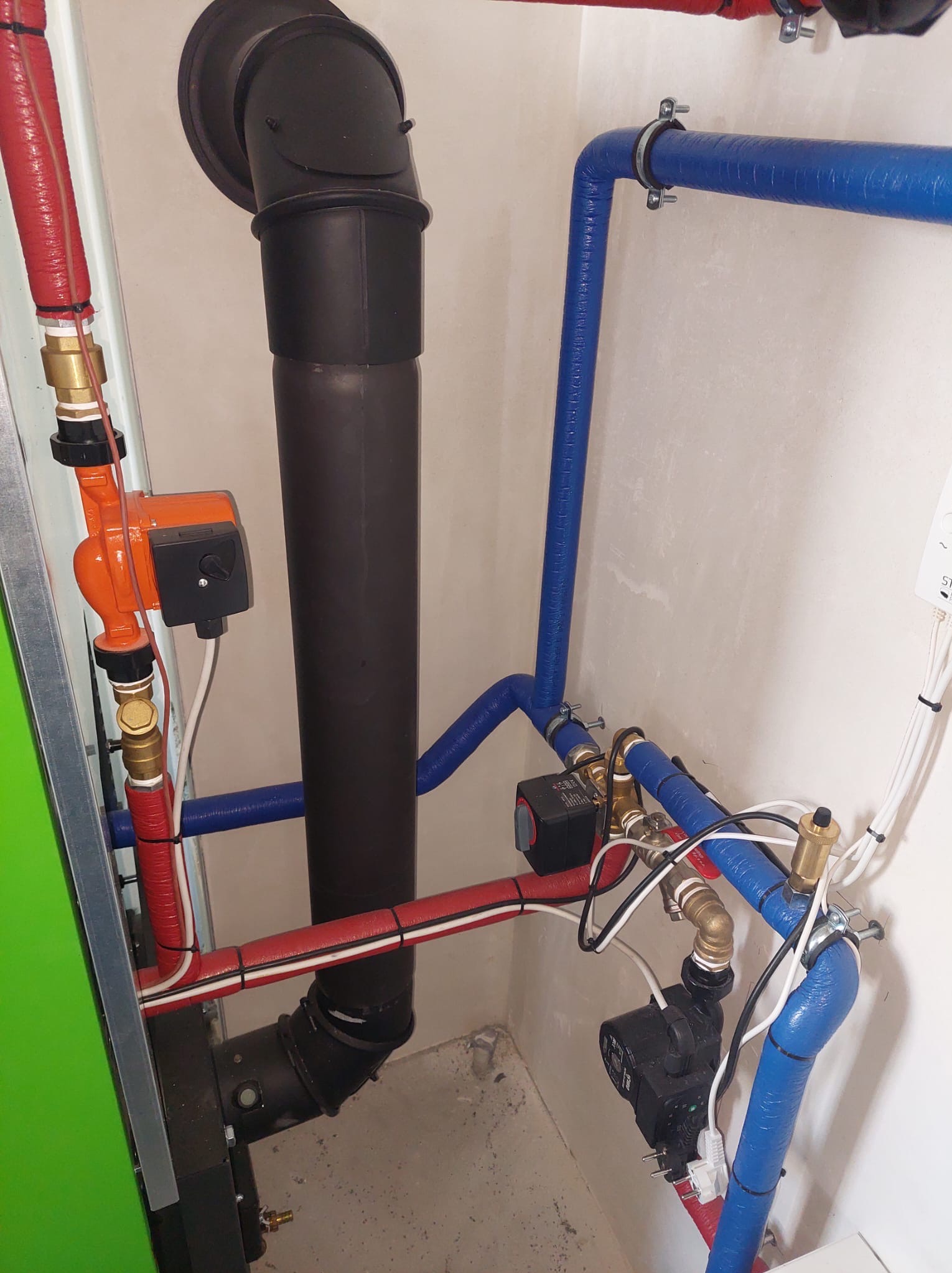 Złożona instalacja grzewcza z rurami w kolorze czerwonym i niebieskim, pompami obiegowymi i czarnym kominem, zamontowana przy ścianie w pomieszczeniu.