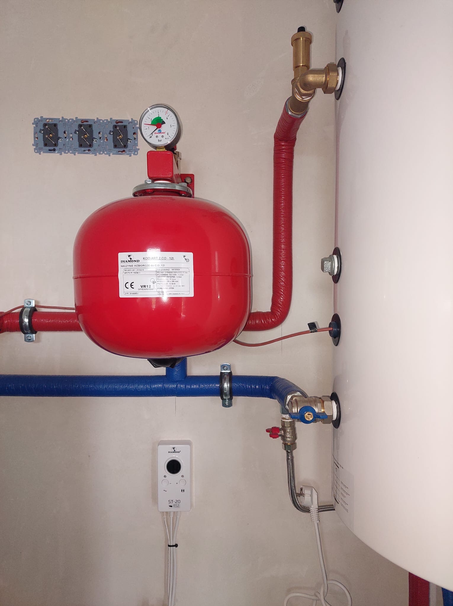 Czerwone naczynie wzbiorcze Diamond z manometrem, podłączone do instalacji CO z niebieskimi i czerwonymi rurami. Widoczny biały bojler i sterownik ST-20 na ścianie.