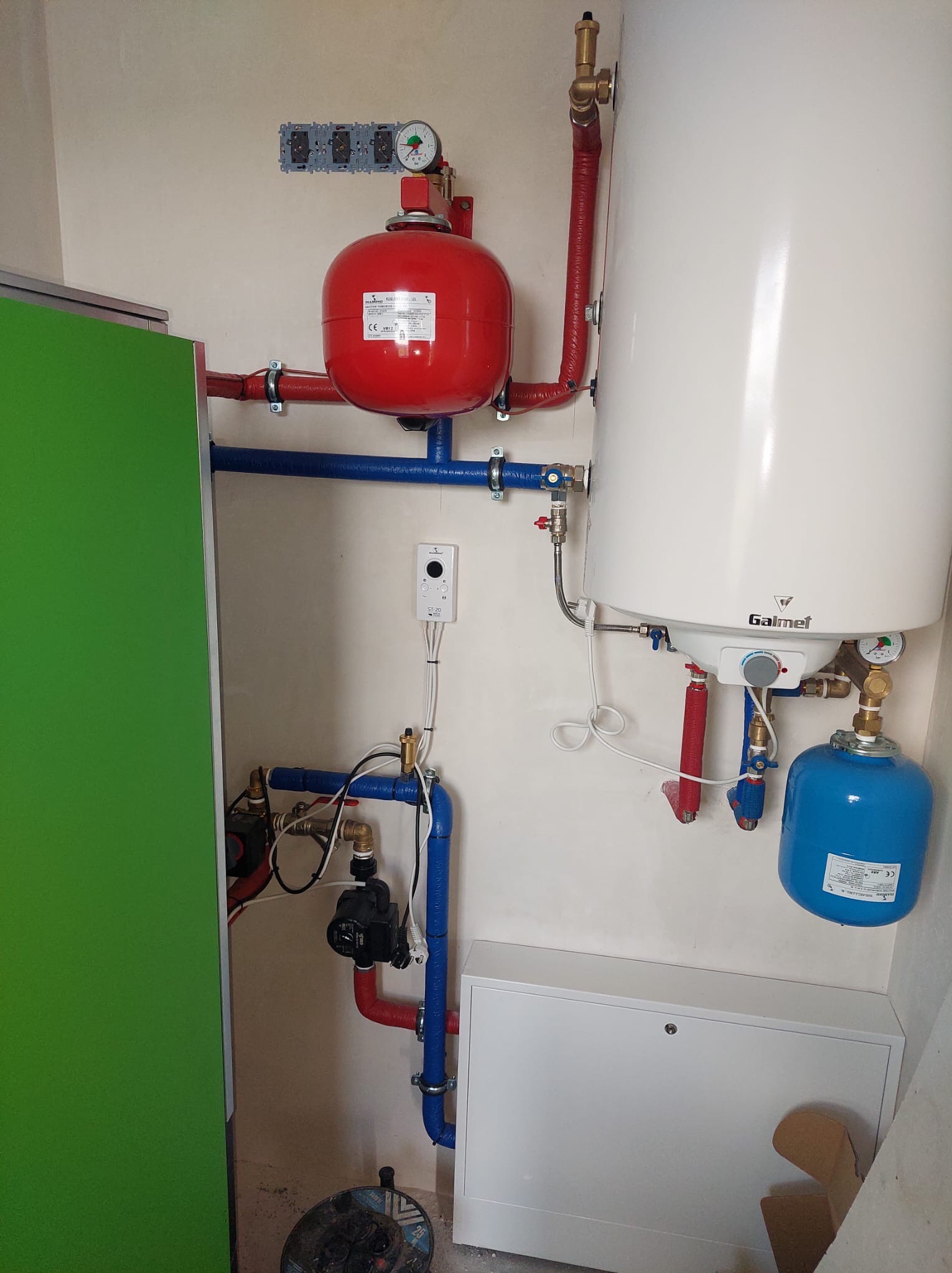 Złożona instalacja grzewcza z czerwonym zbiornikiem wyrównawczym, białym bojlerem Galmet i niebieskimi rurami. Widoczne zawory, manometry i elementy sterujące.