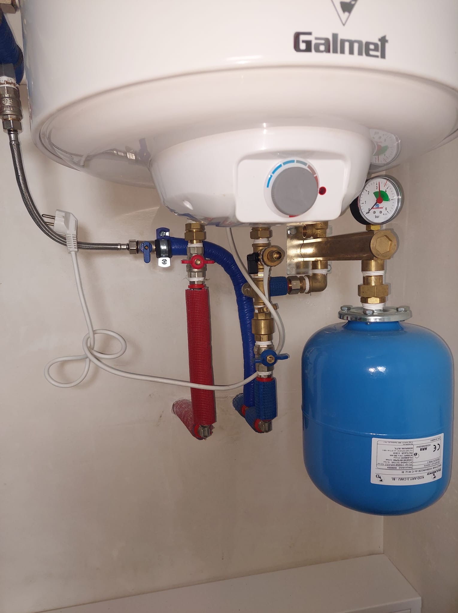 Biały bojler Galmet z niebieskim zbiornikiem wyrównawczym i instalacją hydrauliczną w pomieszczeniu. Widoczne rury w izolacji i manometr.