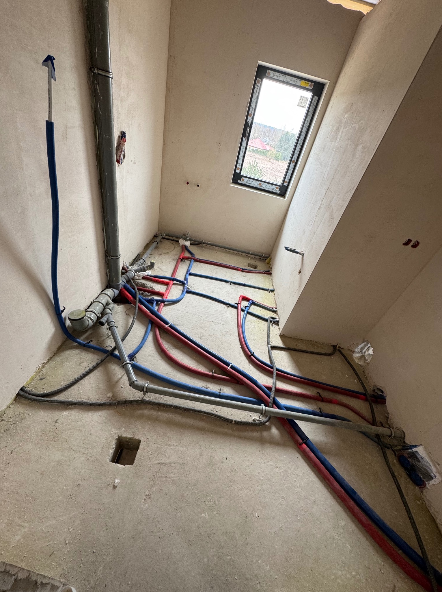 Surowy stan łazienki z oknem dachowym. Widoczne rury instalacji wodnej (czerwone i niebieskie) oraz kanalizacyjne, ułożone na podłodze przed wylaniem posadzki.