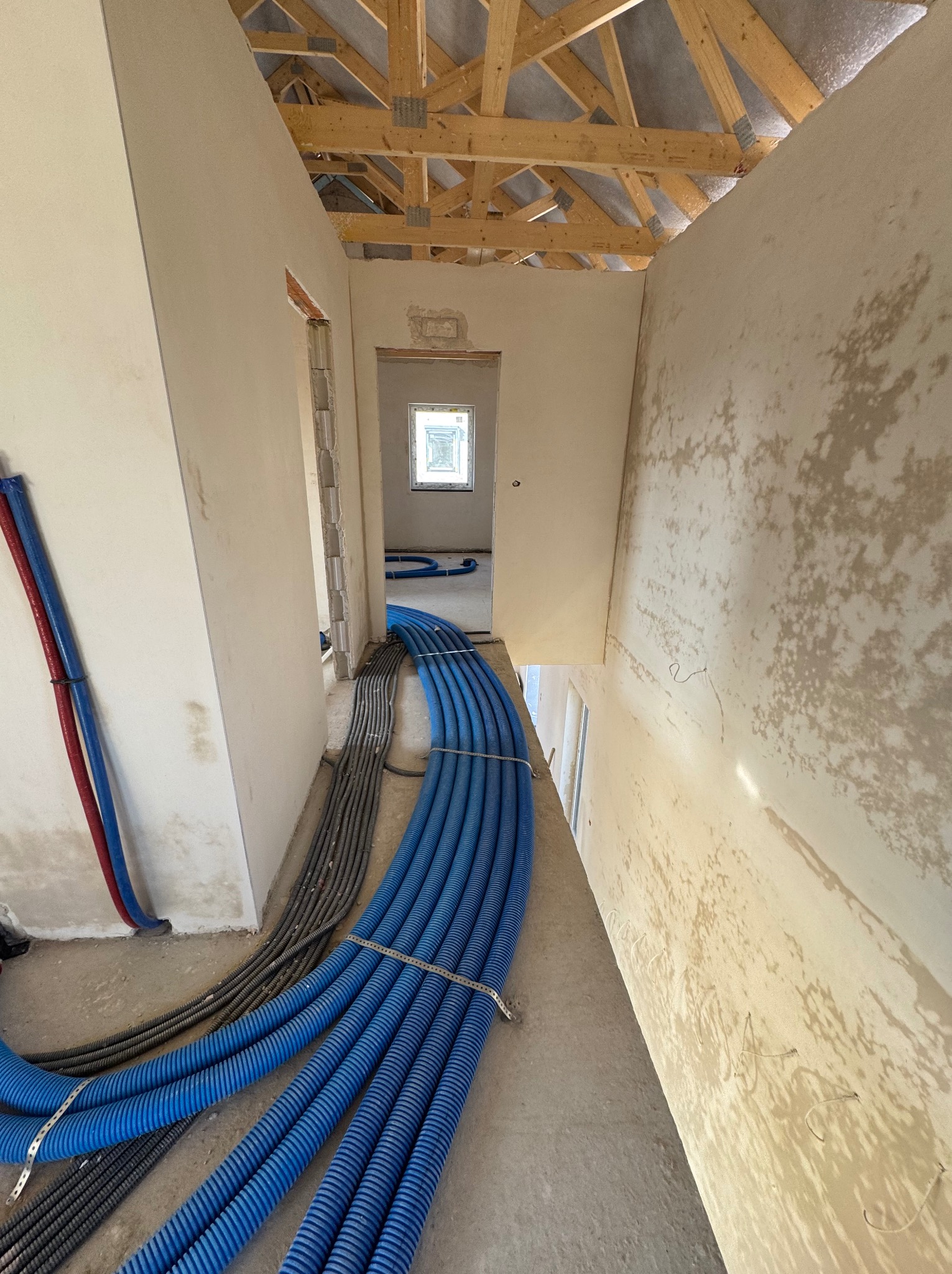 Niebieskie i szare rury instalacyjne ułożone wzdłuż korytarza w surowym stanie, widok z góry, drewniana konstrukcja dachu w tle, ściany w trakcie wykończenia.