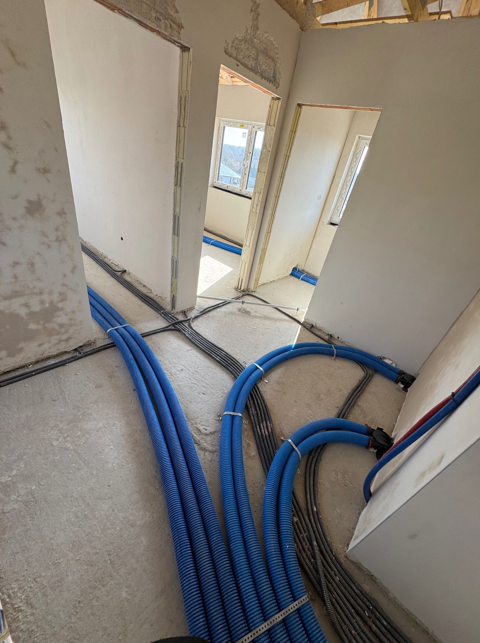 Niebieskie rury instalacyjne na podłodze w surowym wnętrzu budynku we Wrocławiu. Widoczne kable i przygotowane otwory drzwiowe. Instalacja przed wylewką.