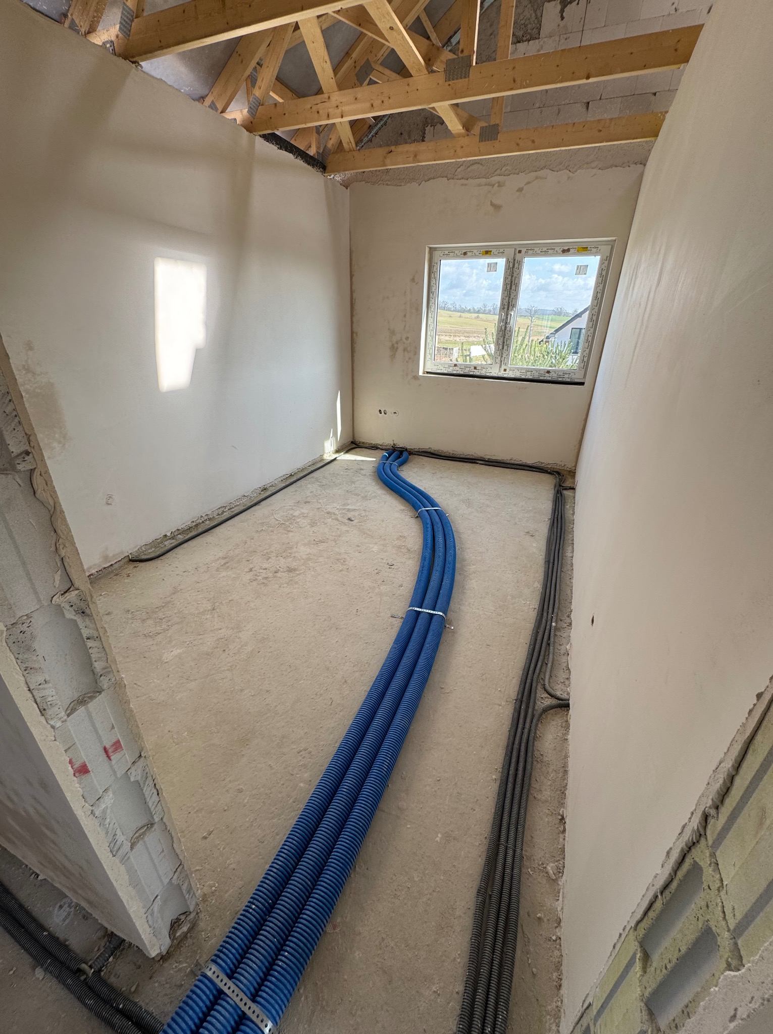 Wnętrze w stanie surowym z ułożonymi, niebieskimi rurami instalacji podłogowej, widoczne okno i surowy strop. Rury przymocowane metalową taśmą.