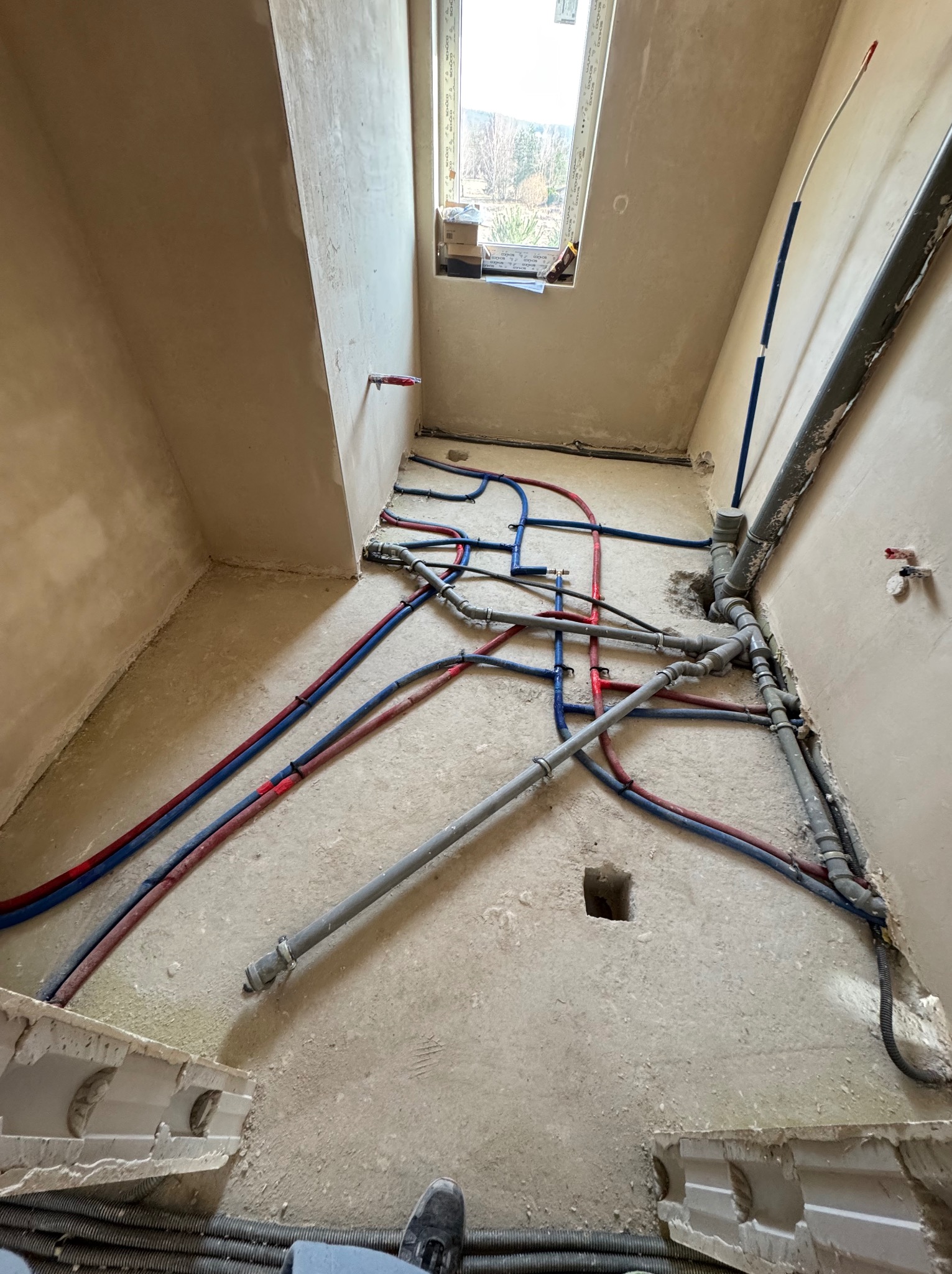 Rozłożone rury instalacji wodnej i kanalizacyjnej w surowym pomieszczeniu z oknem. Widoczne rury czerwone, niebieskie i szare. Częściowo widoczne buty w dolnej części kadru.