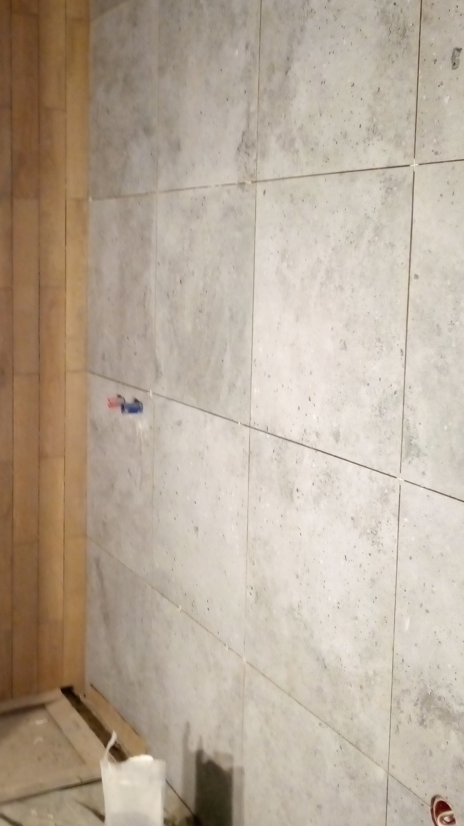 Ściana wyłożona szarymi, kwadratowymi płytkami z widocznymi fugami. Fragment instalacji wodnej z niebiesko-czerwonymi elementami. U dołu widoczny fragment podłogi i wiadro.