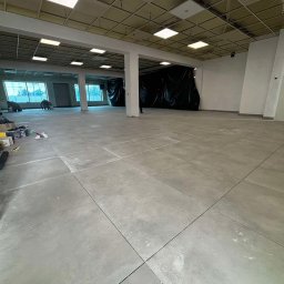 Jakub Witkowski - Wnętrze biura w trakcie prac wykończeniowych: szare płytki podłogowe, białe ściany, sufit podwieszany. Widoczne wiadra z farbą i narzędzia malarskie.