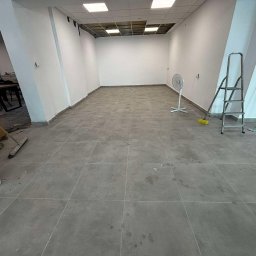 Jakub Witkowski - Wnętrze po remoncie: szare płytki podłogowe, świeżo pomalowane białe ściany, drabina, wentylator i narzędzia malarskie. Widok na sufit z odsłoniętą instalacją.