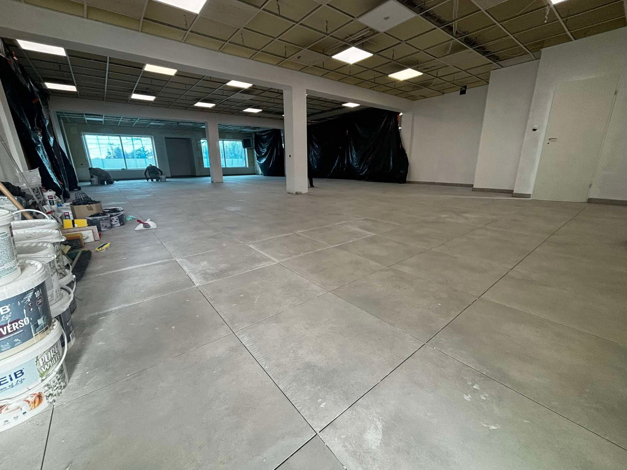 Wnętrze biura w trakcie prac wykończeniowych: szare płytki podłogowe, białe ściany, sufit podwieszany. Widoczne wiadra z farbą i narzędzia malarskie.