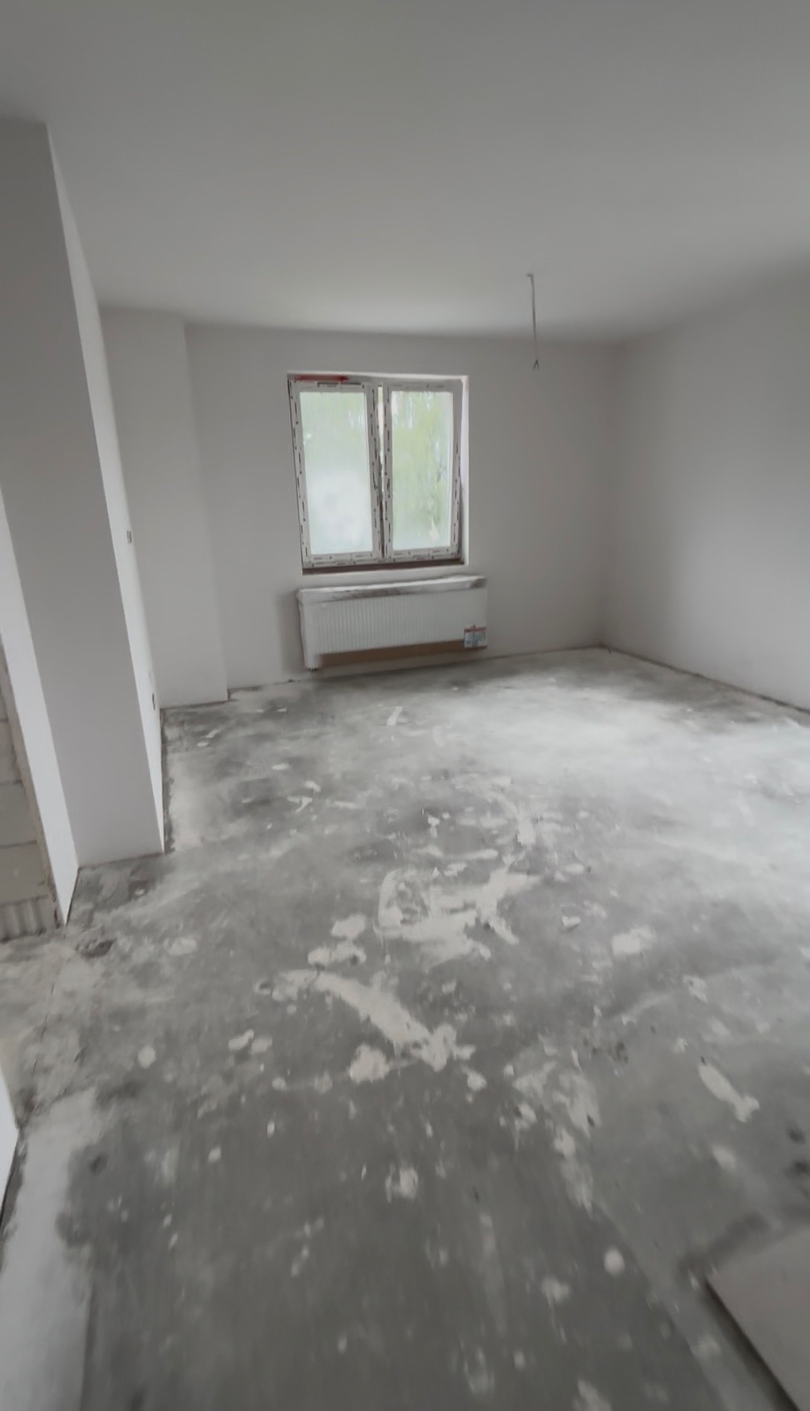 Wnętrze pokoju w trakcie prac wykończeniowych. Widoczne ściany przygotowane do malowania, okno z grzejnikiem i podłoga z pozostałościami po wylewce.