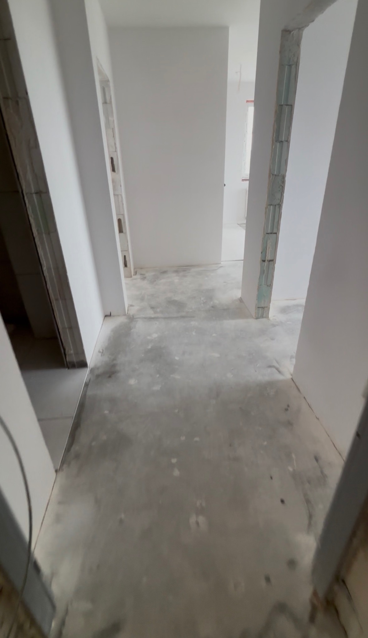 Wnętrze w trakcie prac wykończeniowych: białe ściany, surowa podłoga, widoczne fragmenty konstrukcji ścian. Perspektywa z głębi pomieszczenia.