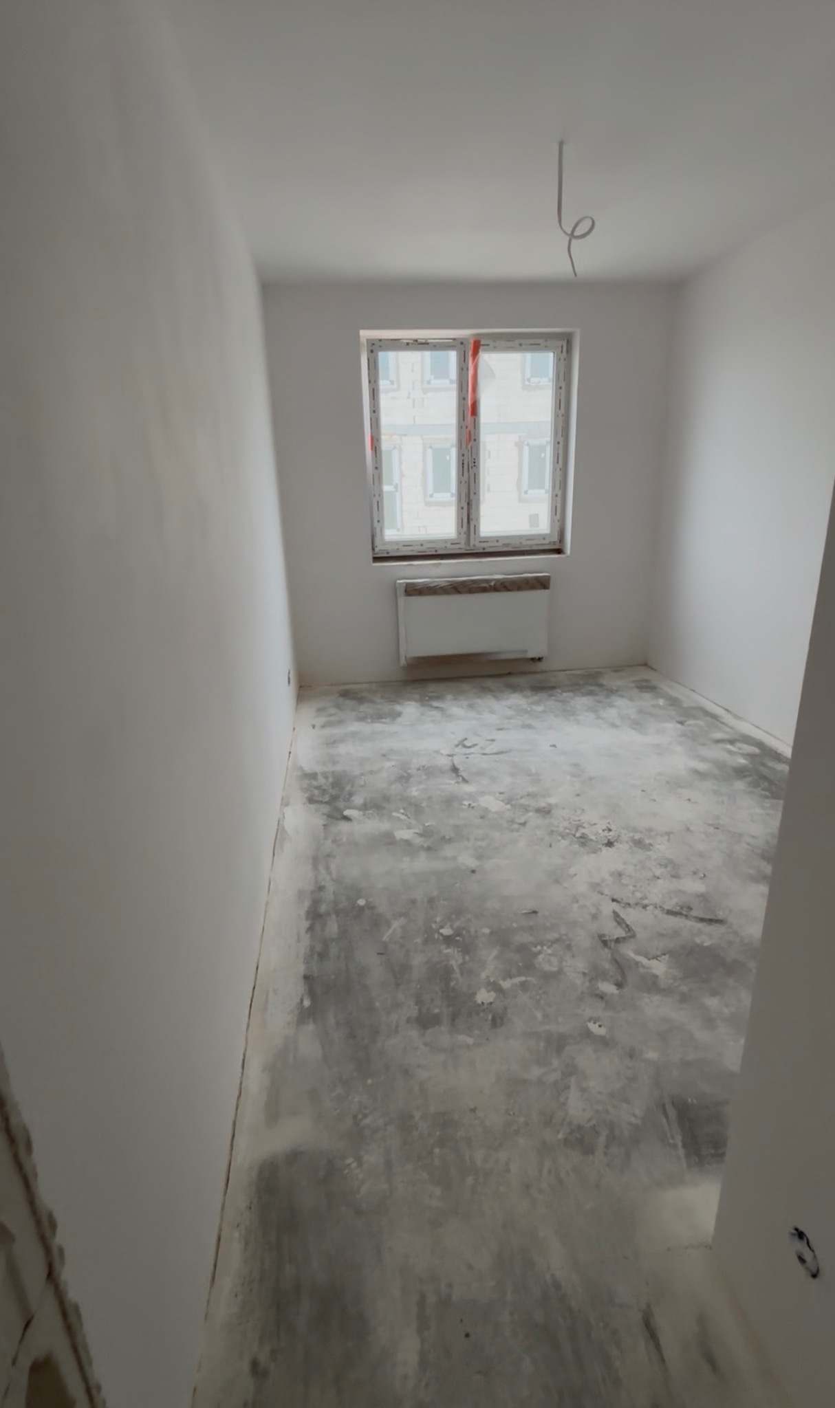 Pokój po wstępnych pracach przygotowawczych: ściany po szpachlowaniu, podłoga z widocznymi śladami prac, okno z grzejnikiem, wystające kable z sufitu.