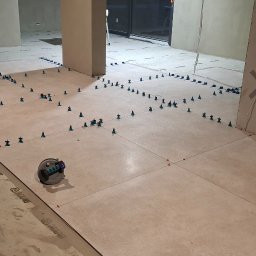 PM-BUD Piotr Michalski - Układanie jasnych płytek podłogowych z użyciem systemu poziomowania. Widoczny laser krzyżowy i przyssawka do przenoszenia płytek. Etap prac wykończeniowych w pomieszczeniu.