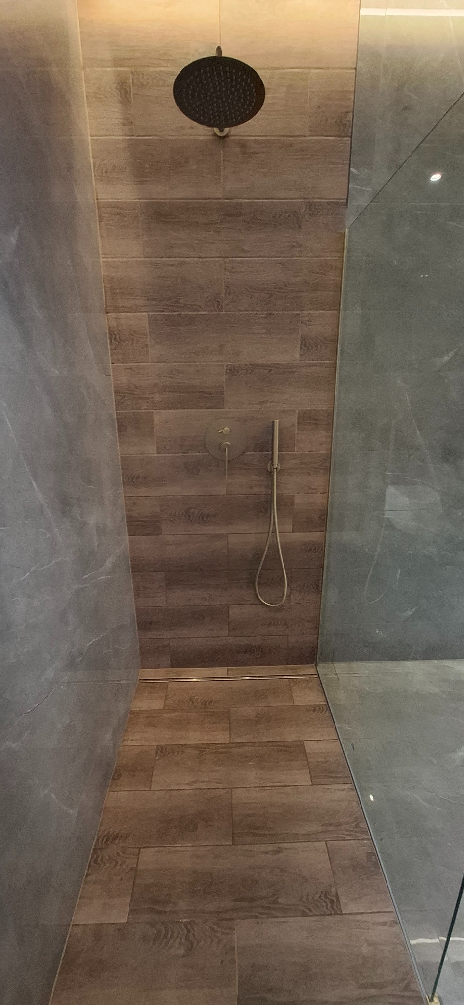 Nowoczesna kabina walk-in z drewnopodobnymi płytkami, odpływem liniowym i baterią podtynkową w odcieniu szczotkowanego złota. Szklana ściana kabiny.