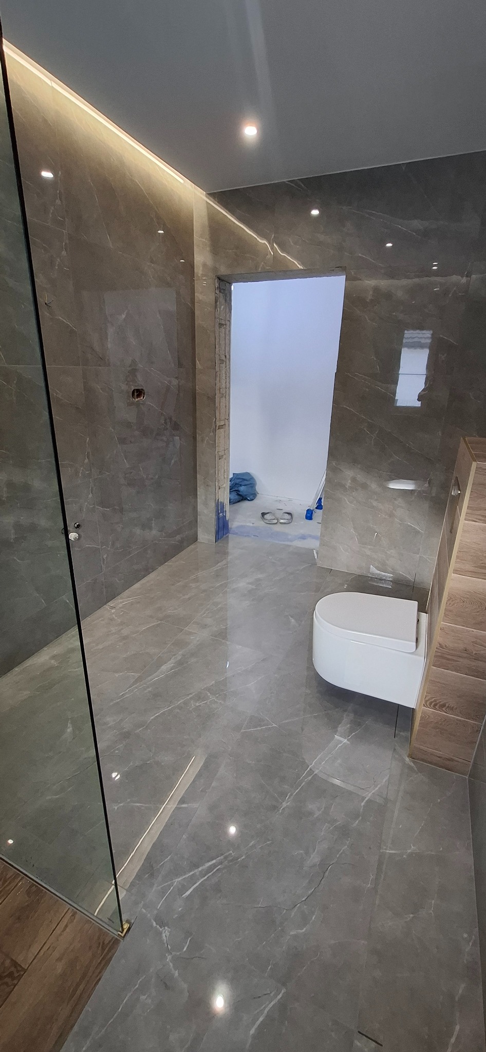Wykończona łazienka z szarymi płytkami imitującymi marmur, podświetleniem LED i częściowo widoczną kabiną prysznicową. W tle widoczne prace wykończeniowe w kolejnym pomieszczeniu.