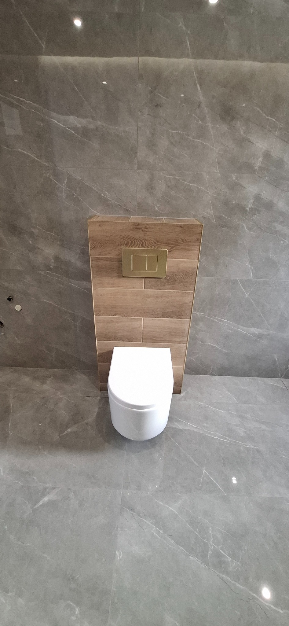 Nowoczesna toaleta podwieszana z drewnianą obudową stelaża i złotym przyciskiem spłukującym, na tle szarych płytek imitujących marmur. Minimalistyczny design.