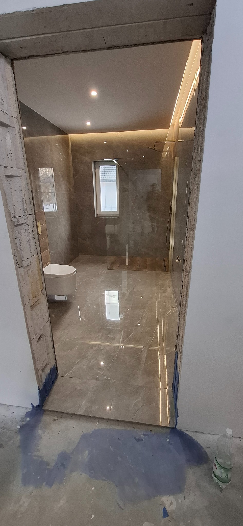 Wykończona łazienka z marmurowymi płytkami, widoczna przez surowy otwór drzwiowy. Nowoczesna kabina prysznicowa i toaleta. Oświetlenie sufitowe.