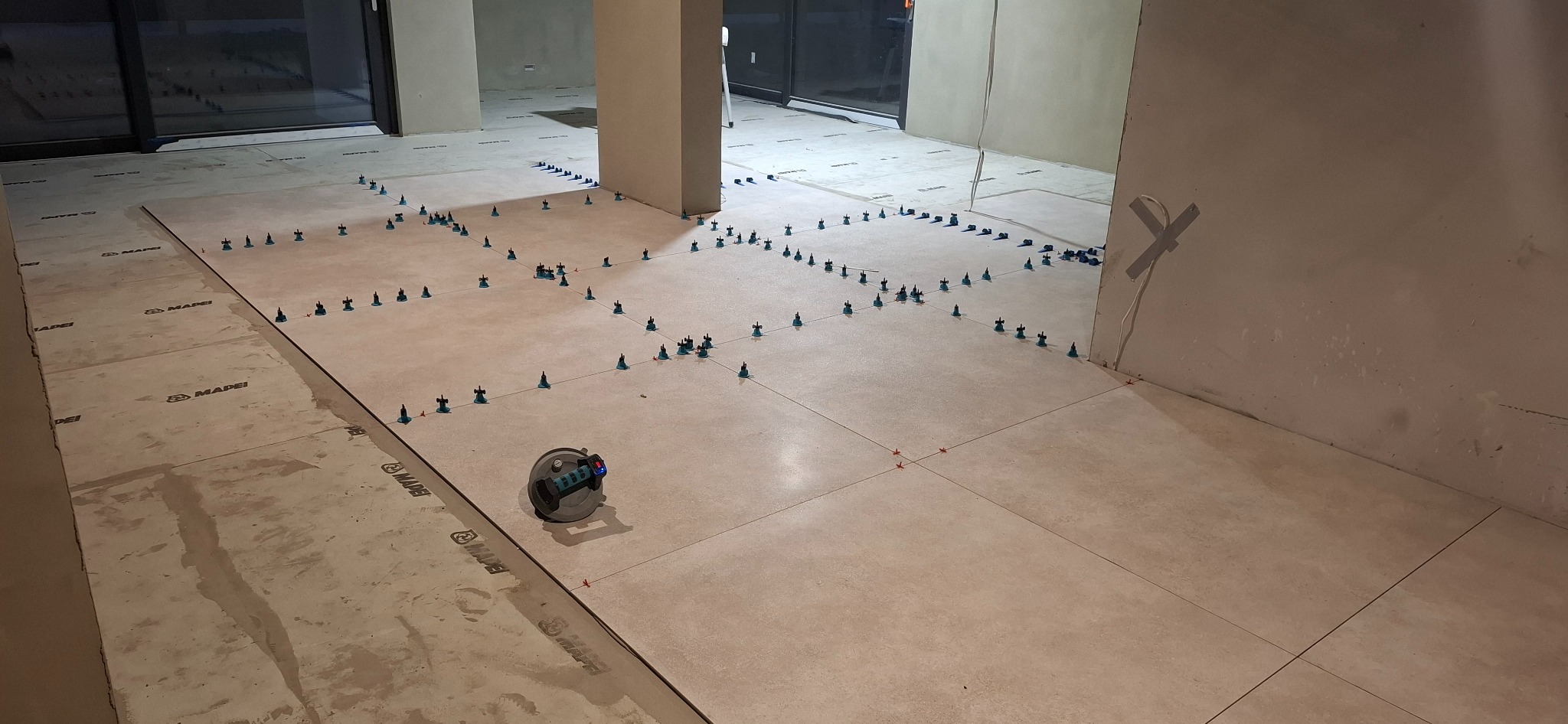 Układanie jasnych płytek podłogowych z użyciem systemu poziomowania. Widoczny laser krzyżowy i przyssawka do przenoszenia płytek. Etap prac wykończeniowych w pomieszczeniu.