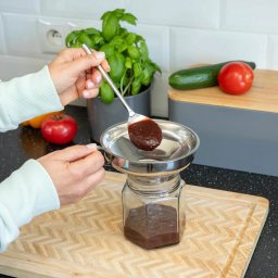 SARA STUDIO - Napełnianie słoika sosem przez metalowy lejek. W tle bazylia, pomidor i ogórek na desce. Jasne wnętrze kuchni z białymi płytkami i czarnym blatem.