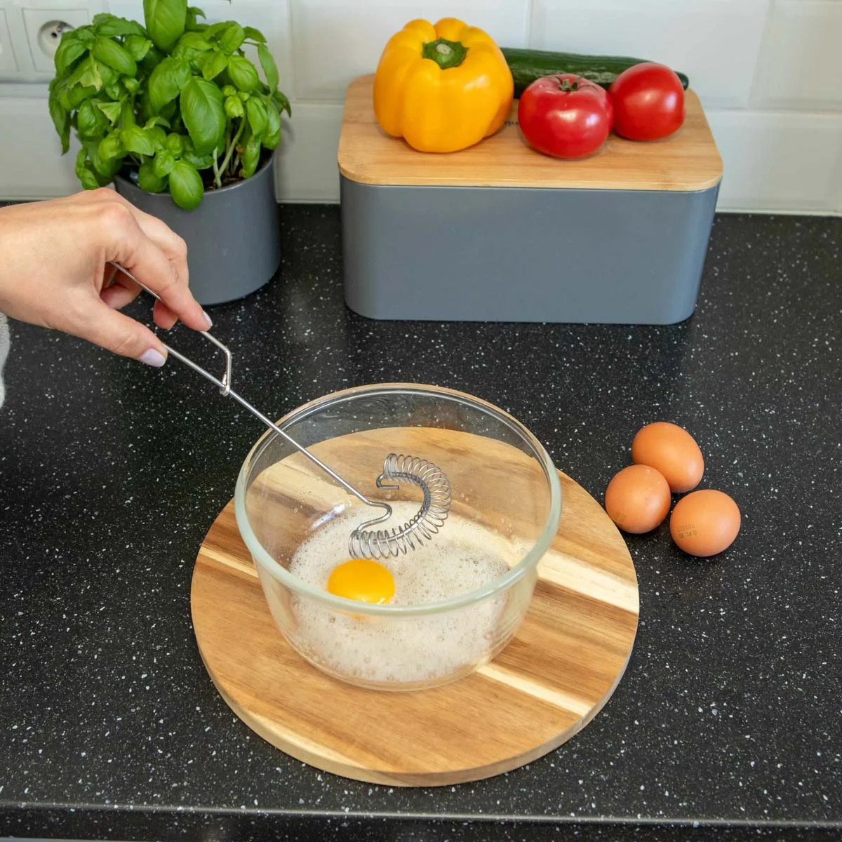 Przygotowanie jajecznicy: ręka ubija jajko w szklanej misce spiralną trzepaczką na drewnianej desce. W tle bazylia, papryka, pomidory i trzy jajka.