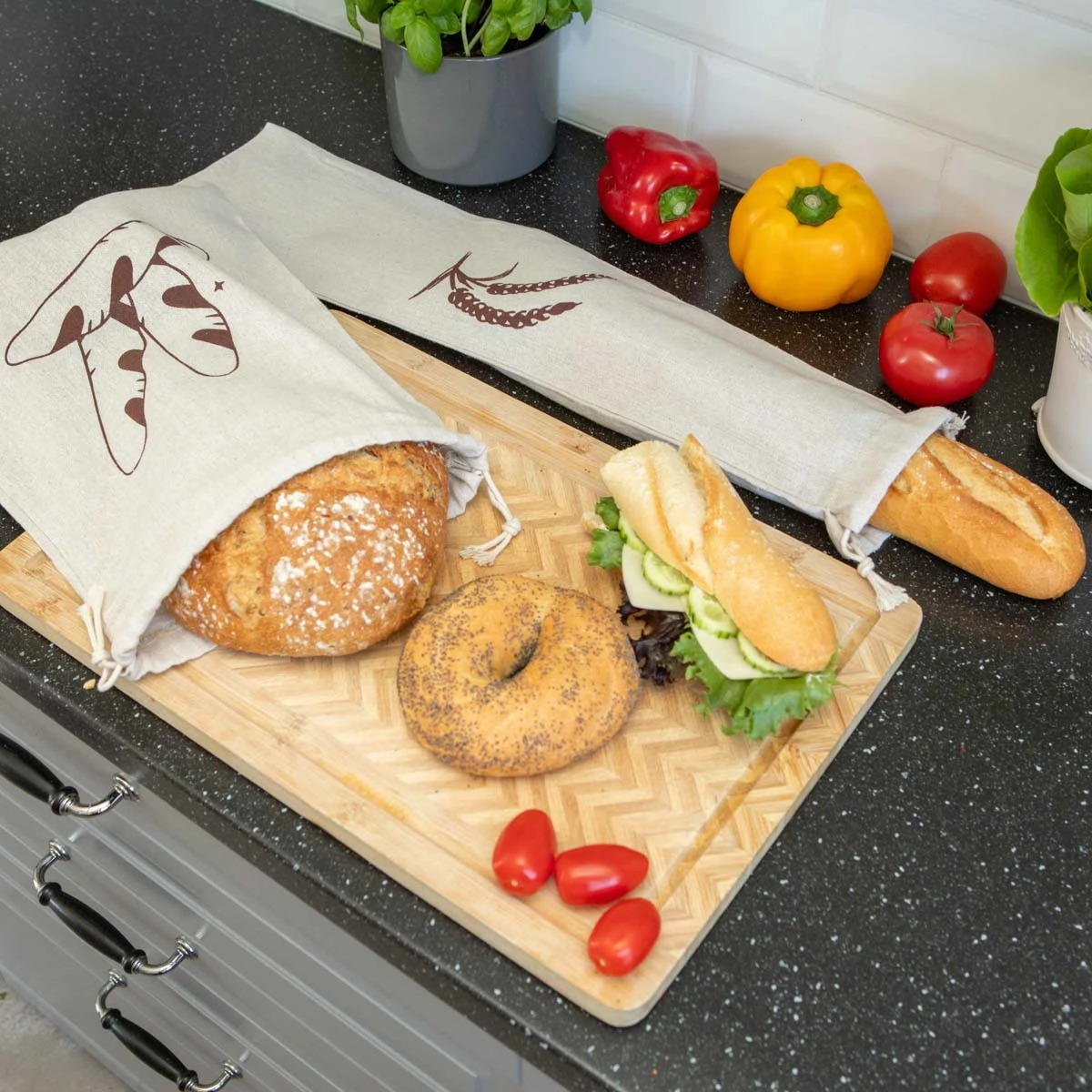 Chleb w lnianych workach z nadrukiem, bajgiel, kanapka na desce do krojenia, kolorowe papryki i pomidory na czarnym blacie kuchennym. Naturalne materiały i zdrowe jedzenie.