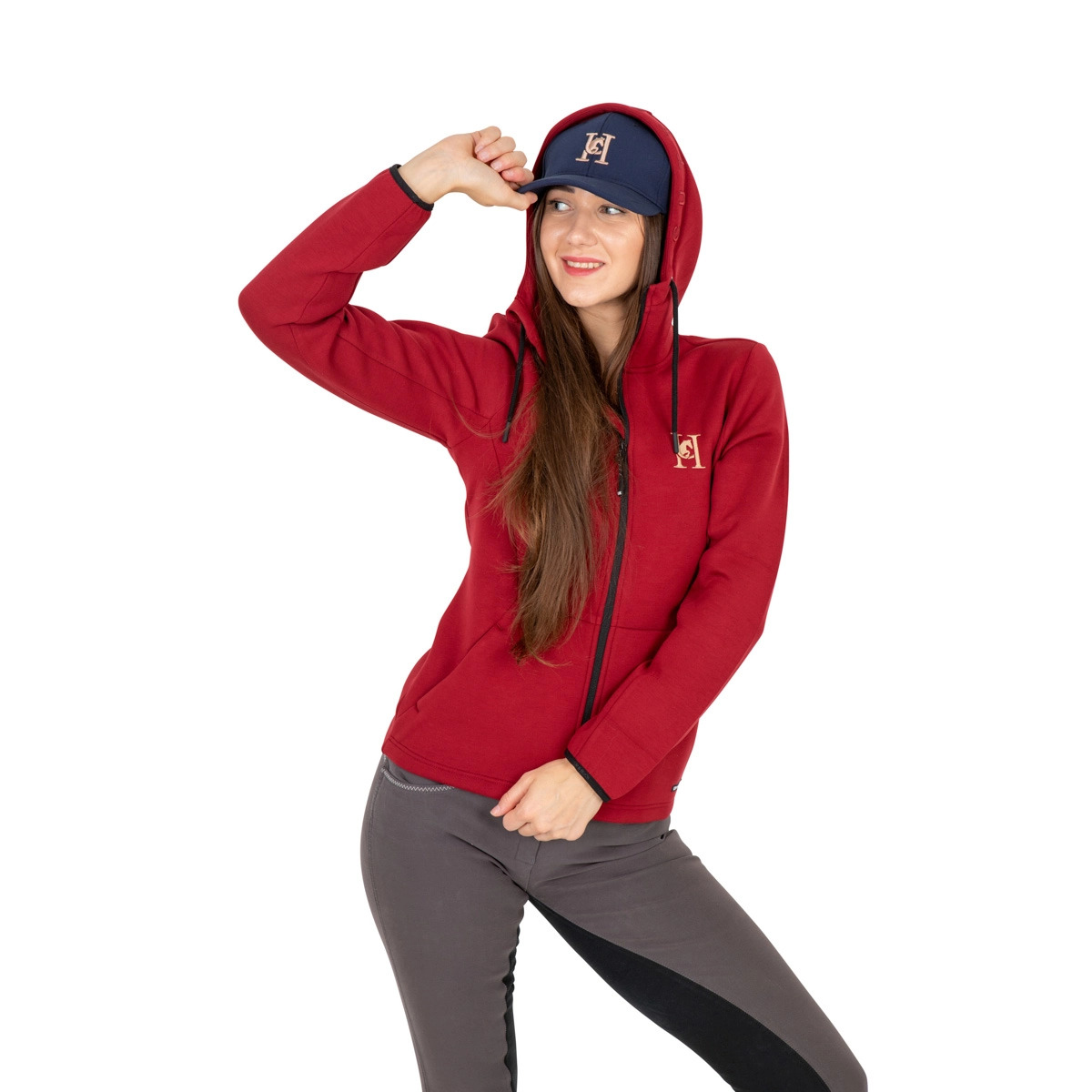 Uśmiechnięta kobieta w czerwonej bluzie z kapturem i granatowej czapce z daszkiem, pozuje na białym tle. Prezentacja odzieży sportowej z logo.