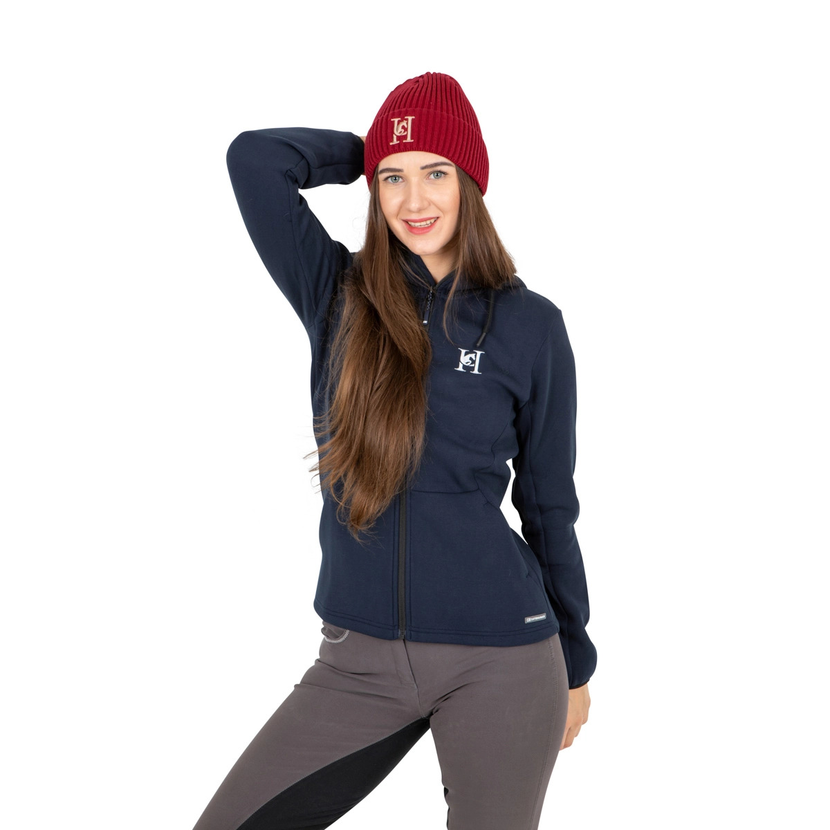 Uśmiechnięta brunetka w bordowej czapce i granatowej bluzie z logo, pozuje na białym tle. Zdjęcie produktowe odzieży jeździeckiej.