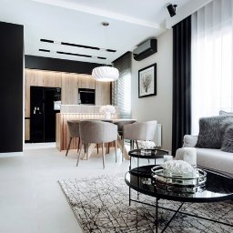 H.P. Agencja Nieruchomości - Jasne, nowoczesne wnętrze: salon z aneksem kuchennym. Drewniane wykończenia, czarne akcenty, okrągły stół z krzesłami i szary narożnik z poduszkami. Minimalistyczny design.