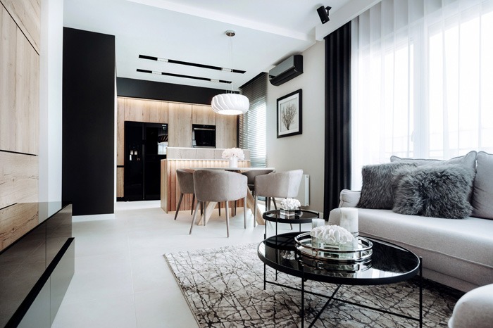 Jasne, nowoczesne wnętrze: salon z aneksem kuchennym. Drewniane wykończenia, czarne akcenty, okrągły stół z krzesłami i szary narożnik z poduszkami. Minimalistyczny design.