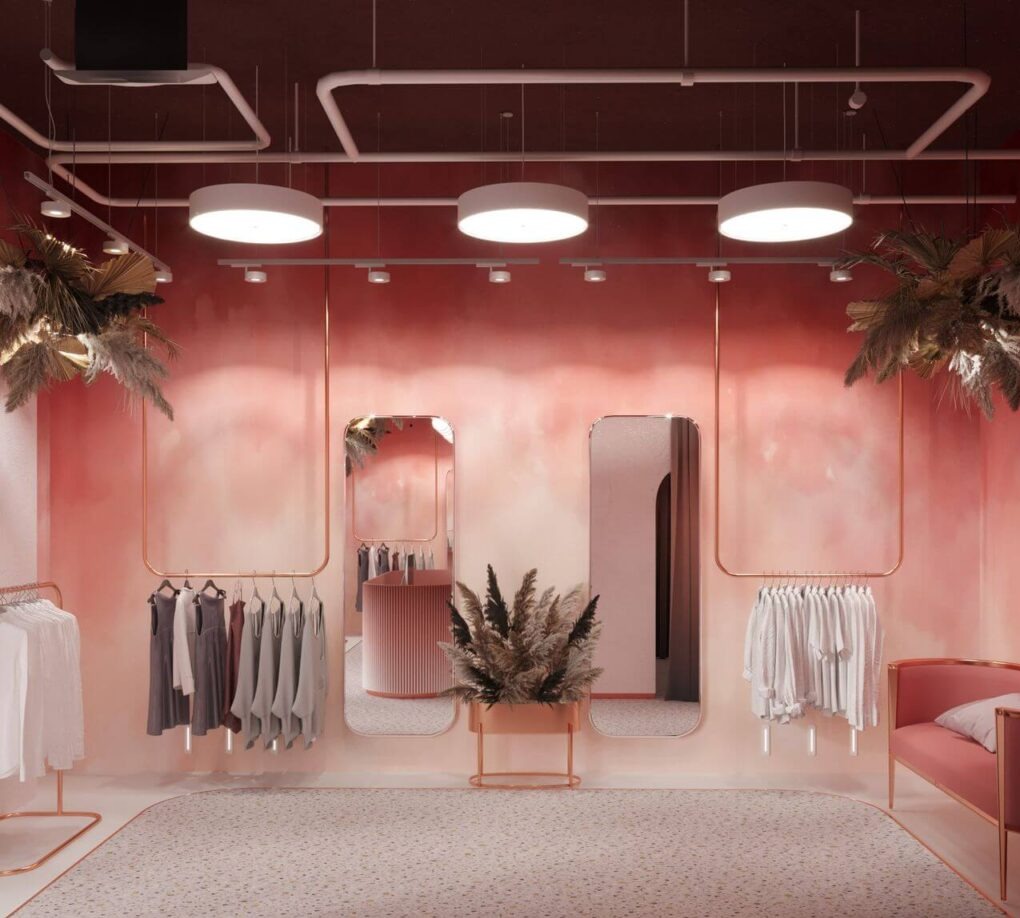 Stylowe wnętrze butiku odzieżowego z różowymi ścianami, złotymi wieszakami i lustrami. Uwagę przykuwają designerskie lampy sufitowe i dekoracje z suszonych traw.