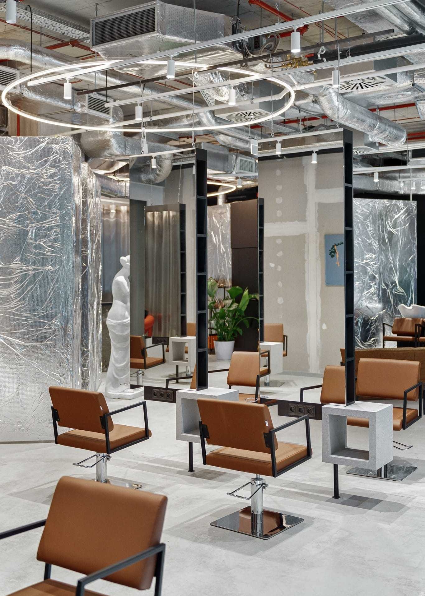 Nowoczesne wnętrze salonu fryzjerskiego z krzesłami w kolorze karmelowym, lustrami i surowym wykończeniem sufitu. Widoczna instalacja oświetleniowa i dekoracyjna statua.