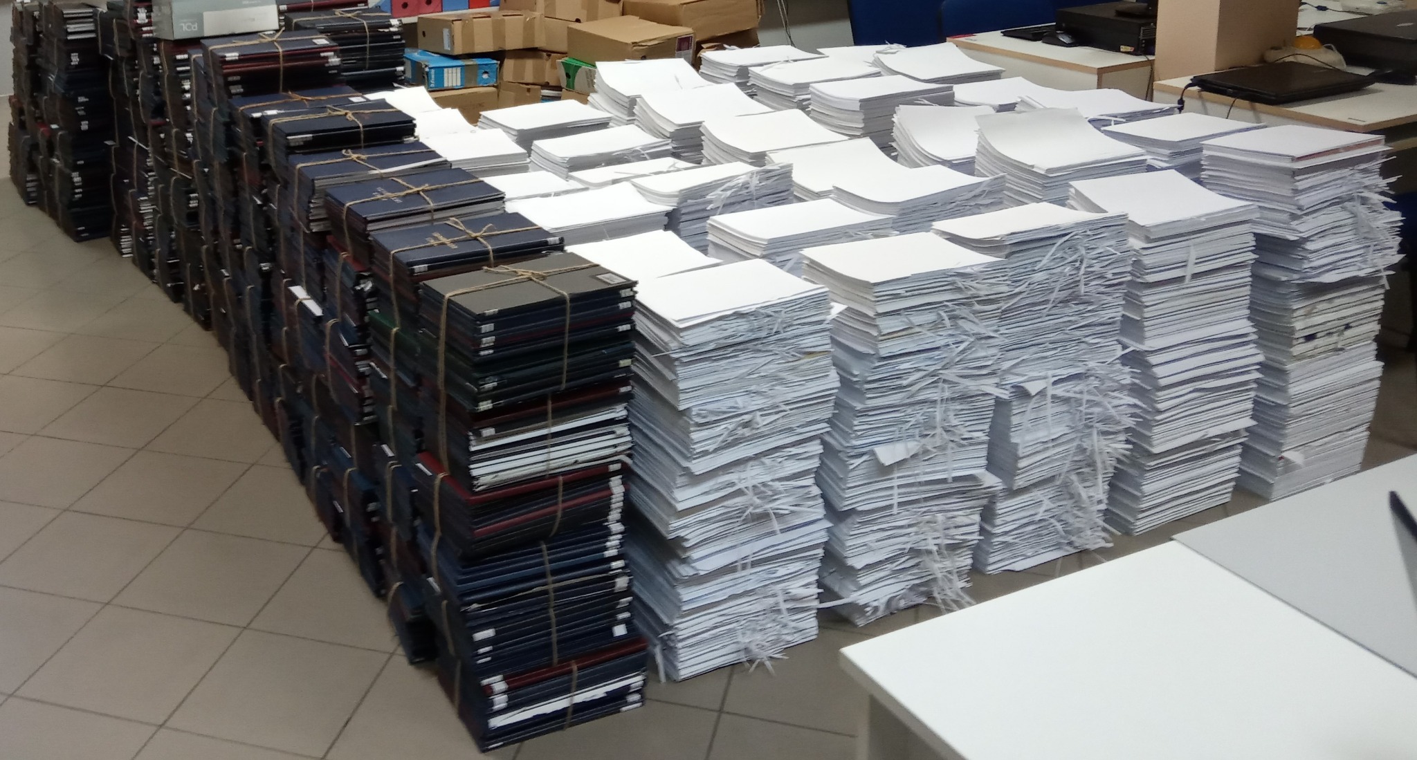 Stosy dokumentów: zarchiwizowane, opasane sznurkiem i pocięte na strzępy, gotowe do utylizacji. Widok z góry, ukazujący skalę pracy.