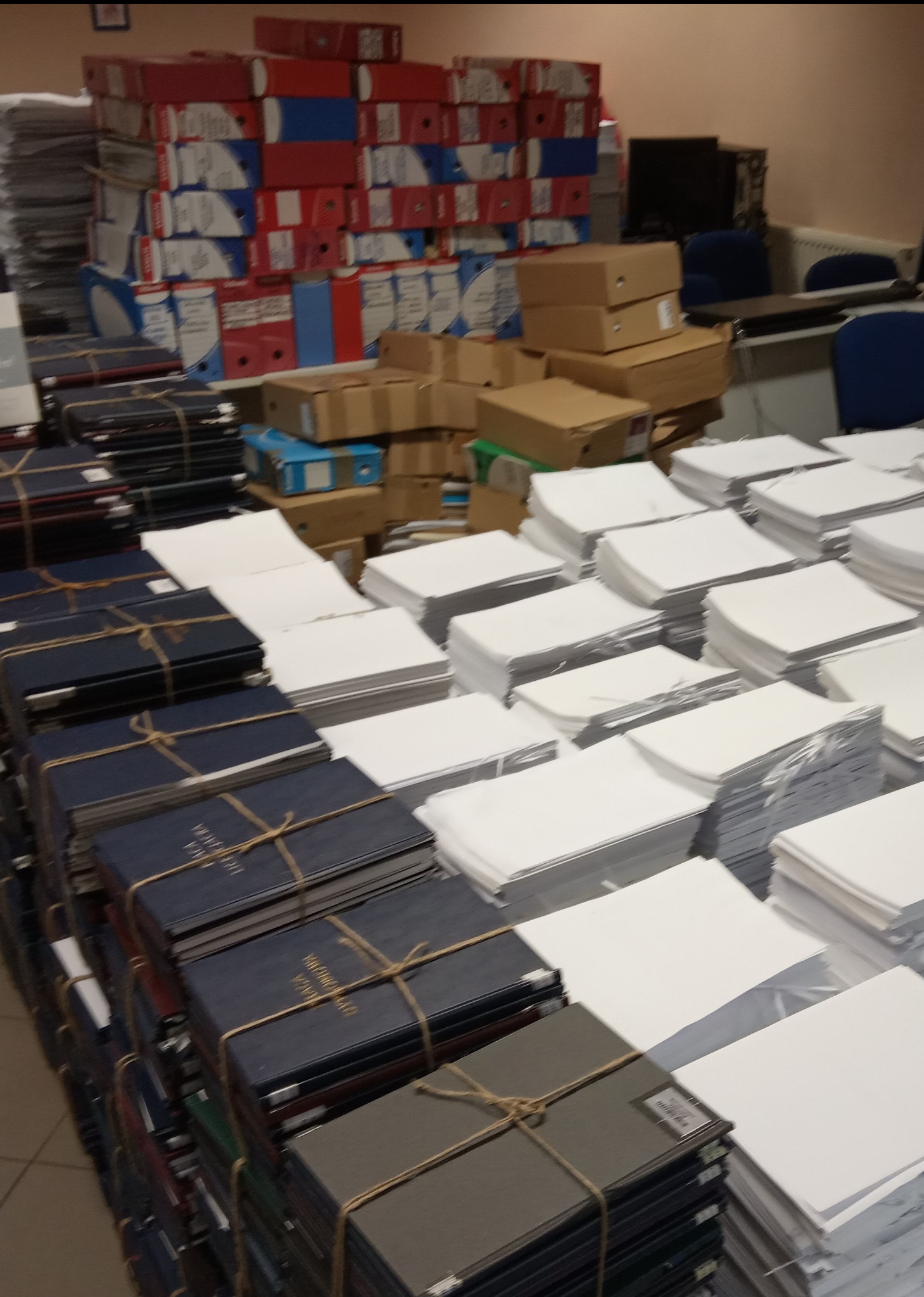 Stosy dokumentów i segregatorów w biurze. Część dokumentów spięta sznurkiem. W tle kartony i regały z segregatorami. Archiwizacja dokumentów w firmie.