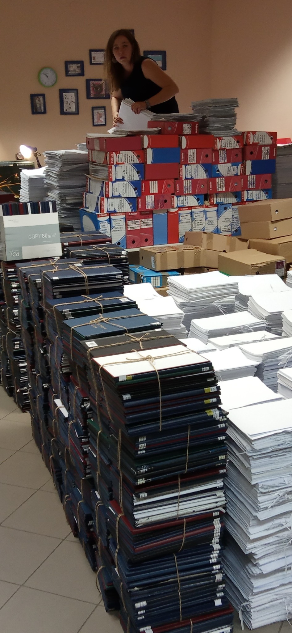 Kobieta układa dokumenty na stosie segregatorów i plikach papieru. Archiwizacja w toku, widoczne segregatory Esselte i paczki papieru 80g/m2.