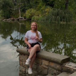 TwinView DroneFilm&Foto - Młoda kobieta siedzi na kamiennym murku z zamkniętymi oczami, w tle jezioro i zieleń. Ubrana w białą koszulkę z motywem serca i wzorzyste spodenki.