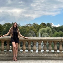 TwinView DroneFilm&Foto - Kobieta w czarnym kombinezonie pozuje na tle kamiennej balustrady z widokiem na fontannę i zieleń parku. Uwagę zwracają wysokie obcasy i długie włosy modelki.