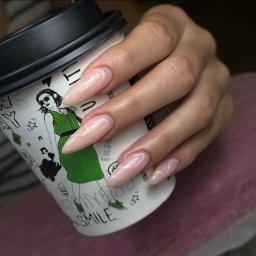 TwinView DroneFilm&Foto - Dłoń z beżowymi paznokciami w kształcie migdała, zdobionymi delikatnym, białym wzorem, trzymająca papierowy kubek z rysunkiem kobiety w zielonej sukience.