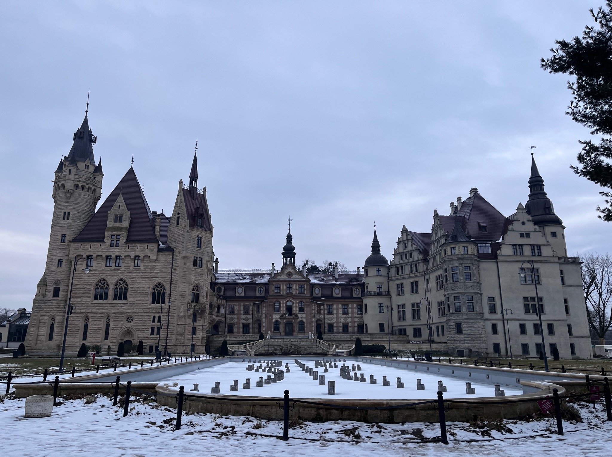 Zimowy kadr na Zamek w Mosznej z fontanną pokrytą śniegiem na pierwszym planie. Pochmurne niebo. Ujęcie z dołu podkreśla monumentalność architektury.