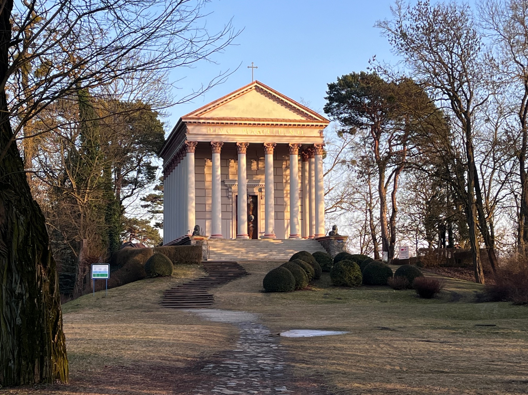 Klasycystyczna świątynia z kolumnami, schodami i krzyżem na szczycie, otoczona drzewami i krzewami w parku, sfotografowana w ciepłym świetle wczesnego poranka.