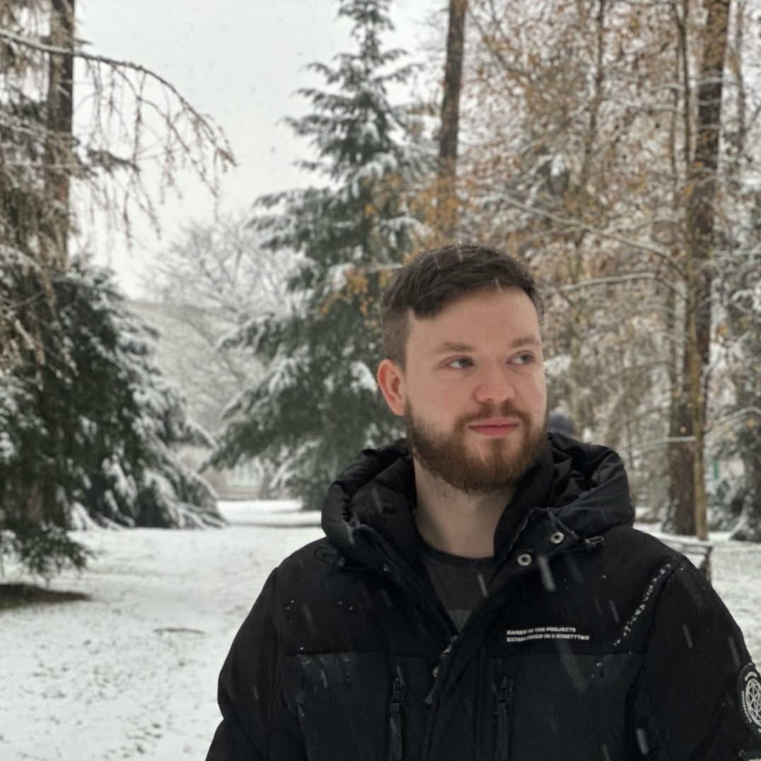 Młody mężczyzna z brodą w czarnej kurtce na tle zimowego parku podczas opadów śniegu, spoglądający w bok.