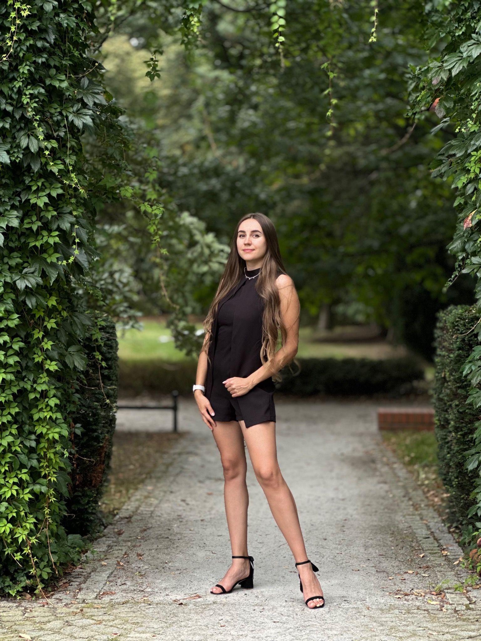 Portret kobiety w czarnym kombinezonie na tle zielonego parku. Ujęcie z dołu podkreśla sylwetkę modelki, a naturalne światło dodaje świeżości.