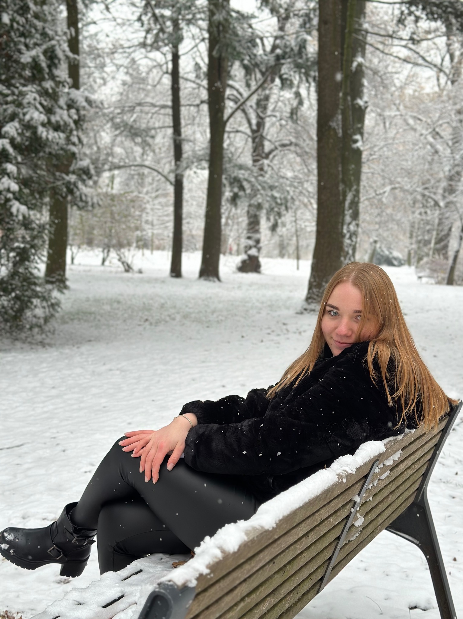 Młoda kobieta z długimi blond włosami siedzi na ławce w parku pokrytym śniegiem, ubrana w czarne futro i legginsy, z delikatnym uśmiechem.