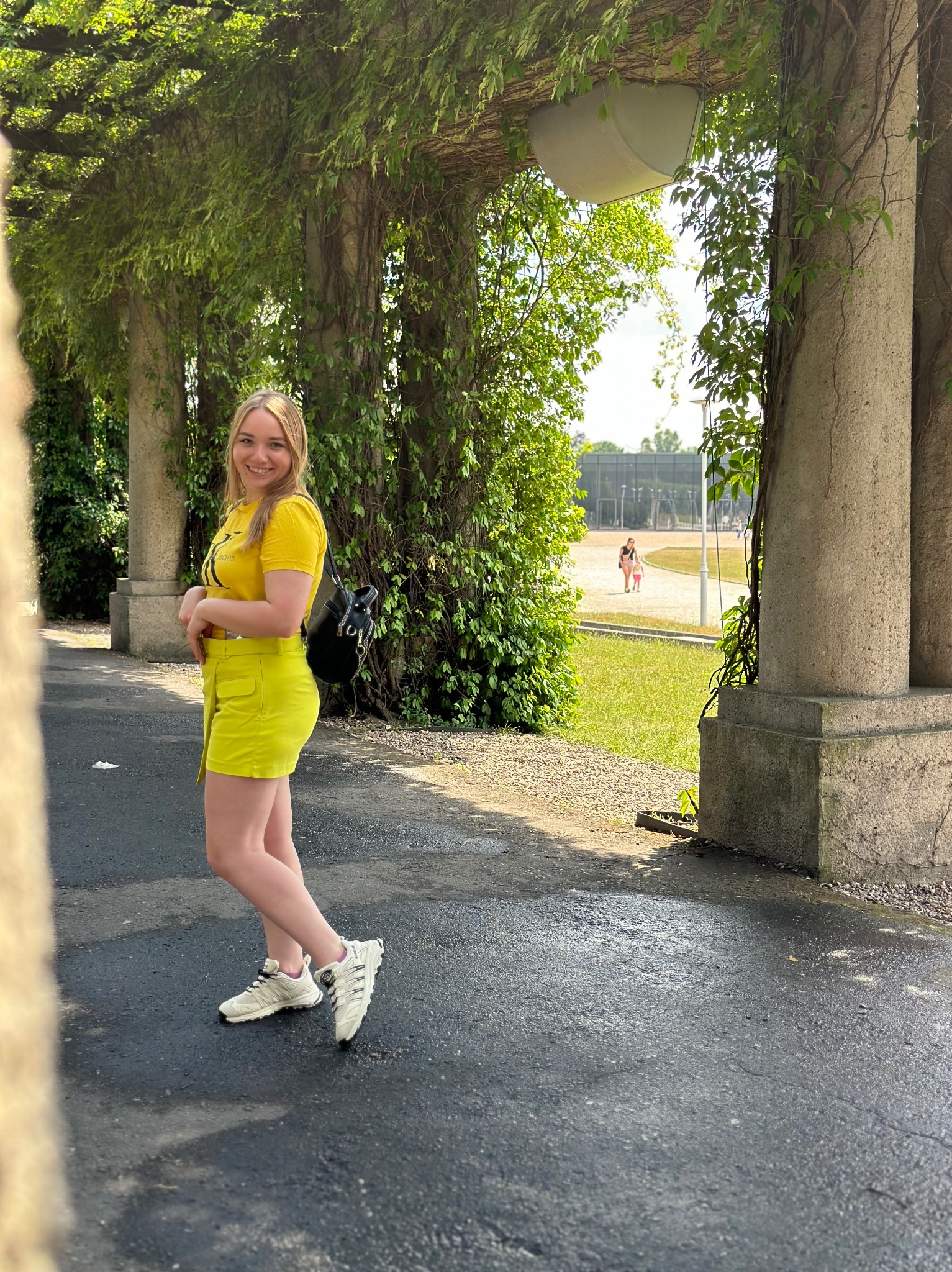 Uśmiechnięta blondynka w neonowożółtym komplecie, pozuje na tle kolumnady porośniętej bluszczem, z widokiem na park i spacerujących ludzi w oddali.