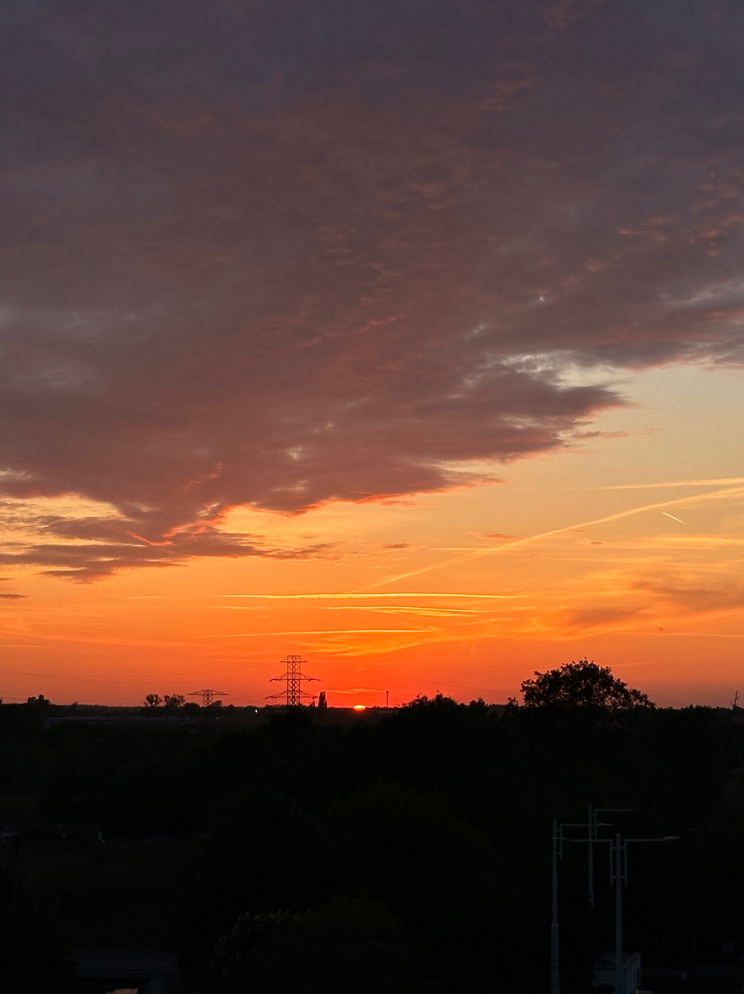 Pomarańczowy zachód słońca nad miastem z widocznymi liniami smug po samolotach i sylwetkami drzew oraz słupów wysokiego napięcia na horyzoncie.