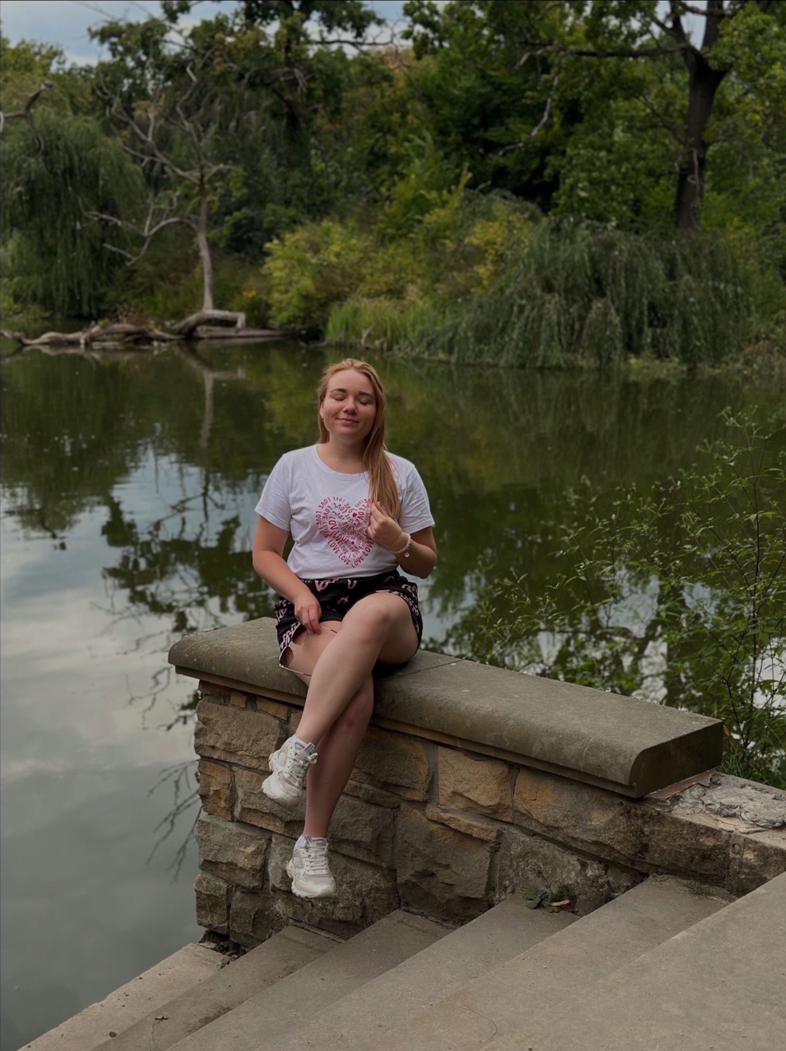 Młoda kobieta siedzi na kamiennym murku z zamkniętymi oczami, w tle jezioro i zieleń. Ubrana w białą koszulkę z motywem serca i wzorzyste spodenki.