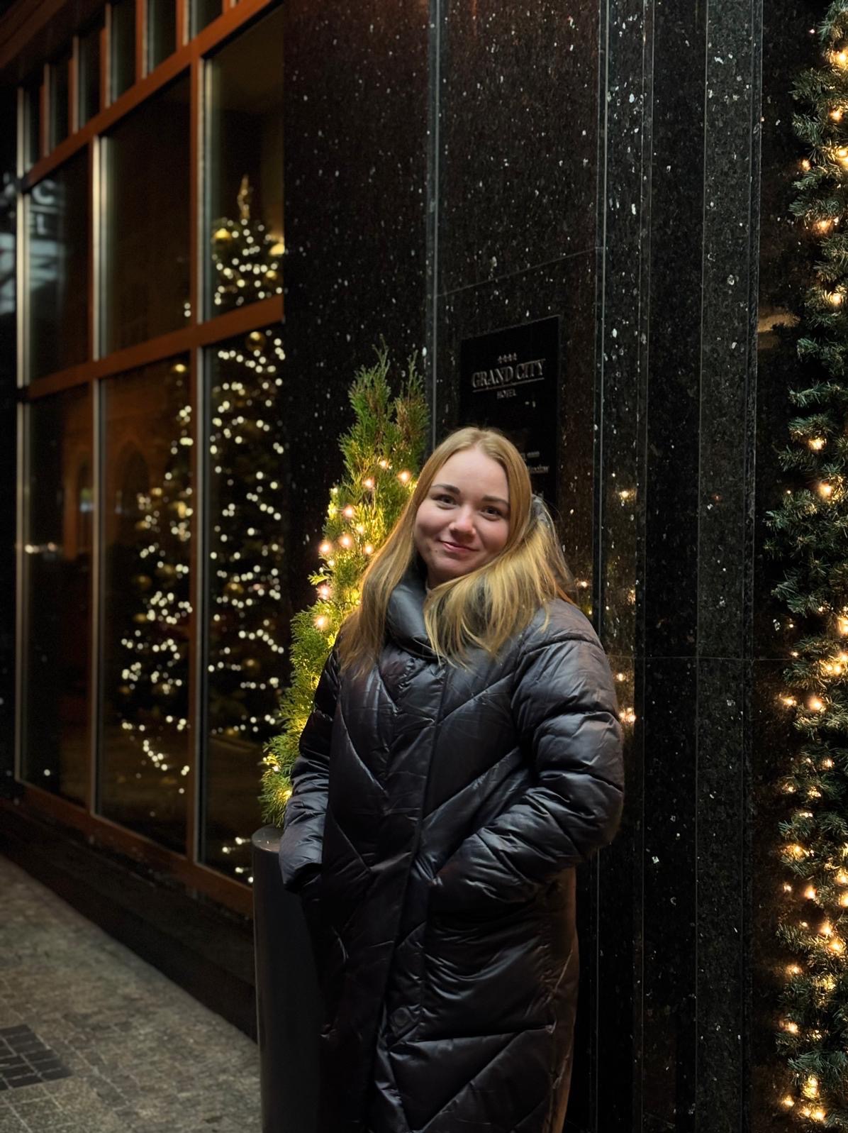 Portret kobiety w czarnej kurtce na tle świątecznej dekoracji hotelu Grand City. W tle okno z choinką i światełka.