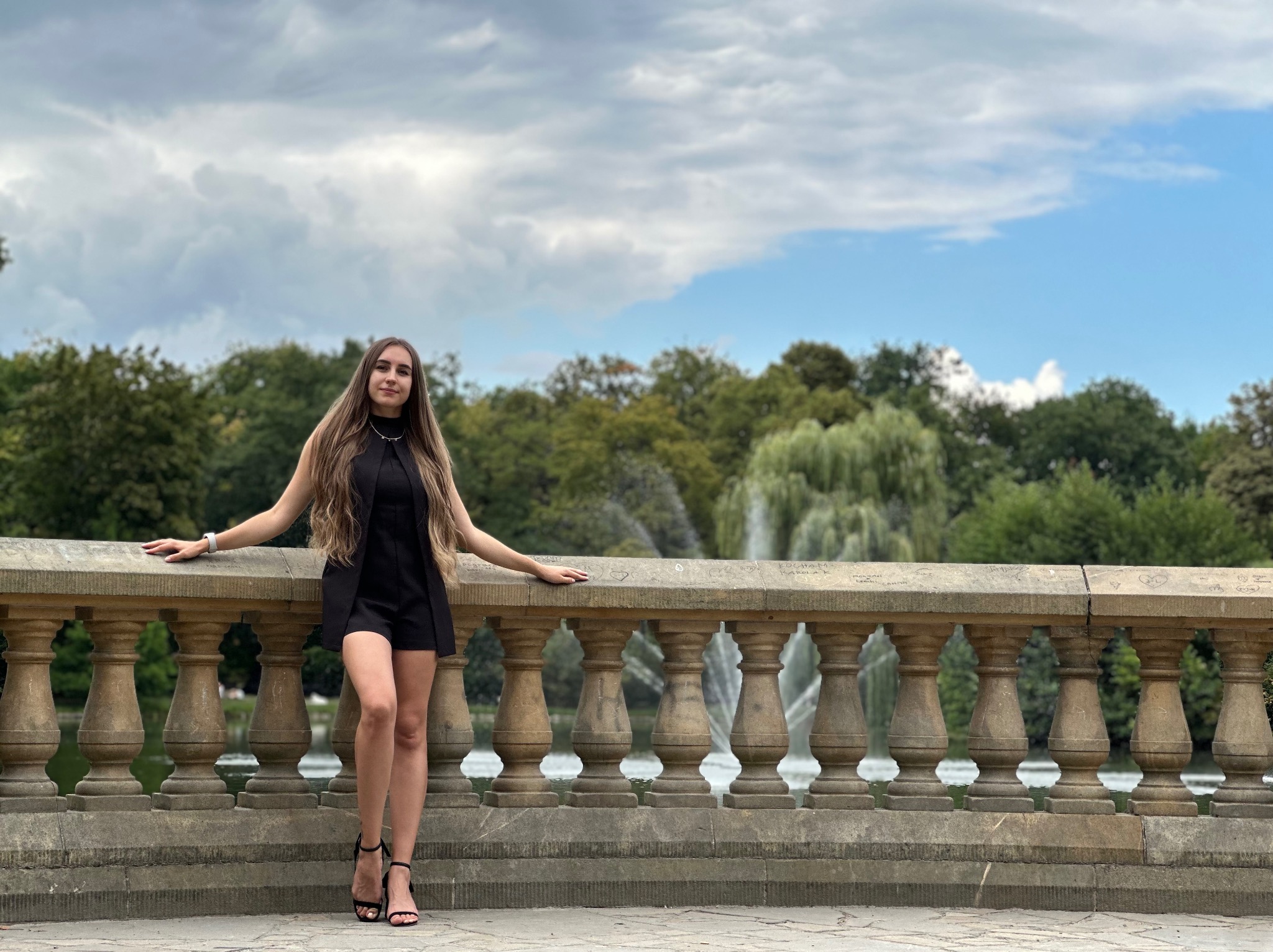 Kobieta w czarnym kombinezonie pozuje na tle kamiennej balustrady z widokiem na fontannę i zieleń parku. Uwagę zwracają wysokie obcasy i długie włosy modelki.