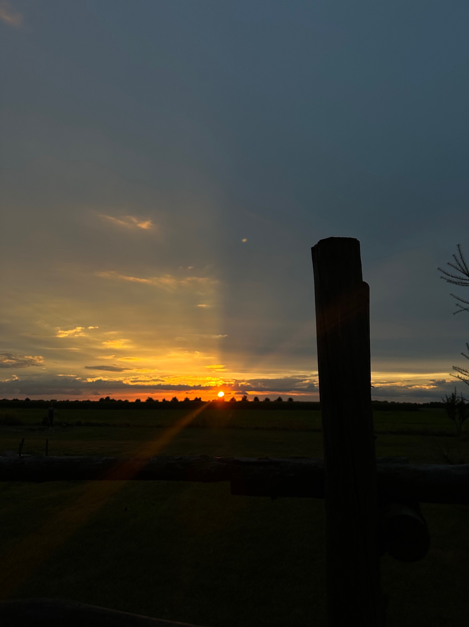 Zachód słońca nad polami, widziany przez drewniane ogrodzenie. Ciepłe barwy nieba kontrastują z ciemnym pierwszym planem. Promienie słońca przebijają się przez chmury.