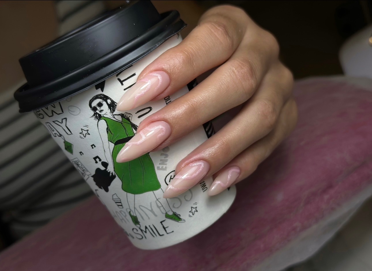 Dłoń z beżowymi paznokciami w kształcie migdała, zdobionymi delikatnym, białym wzorem, trzymająca papierowy kubek z rysunkiem kobiety w zielonej sukience.