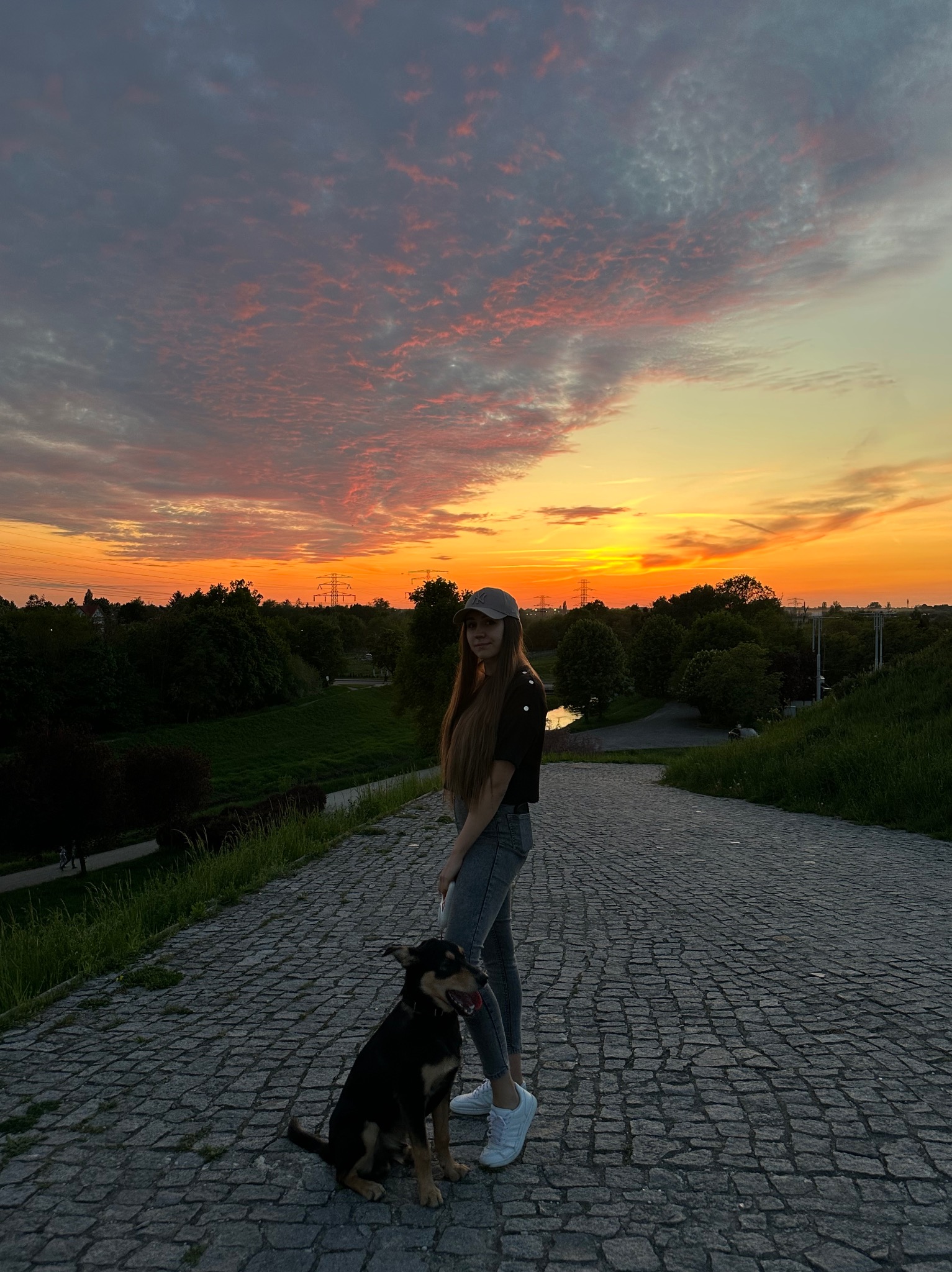 Młoda kobieta z psem na brukowanej ścieżce o zmierzchu. Pomarańczowe niebo, drzewa w tle. Ujęcie z dołu, podkreślające perspektywę i rozległość krajobrazu.